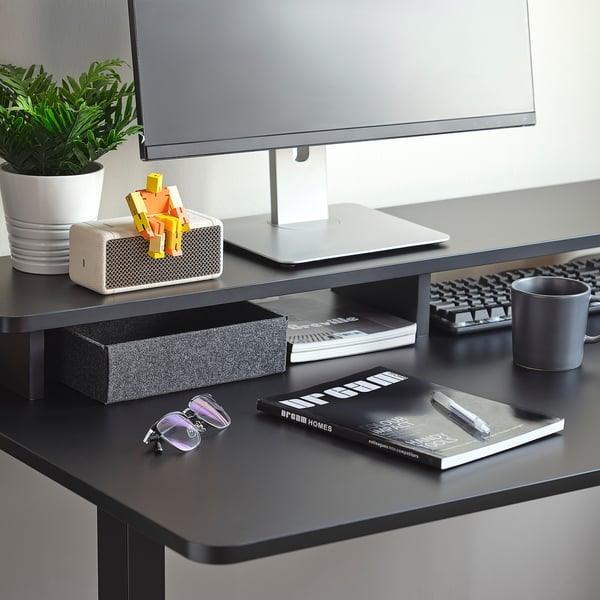 Modern desk with monitor stand, books, cup, and plant. White speaker with colourful blocks on top.