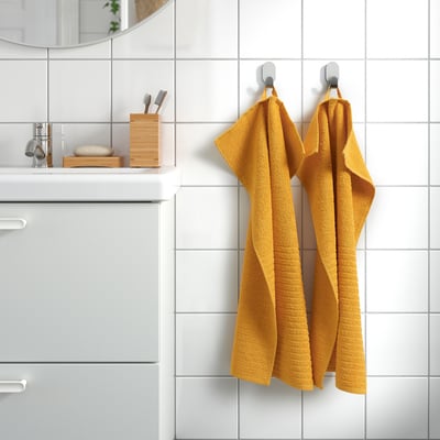 Yellow VÅGSJÖN bath towels hang neatly on hooks, next to sink with white cabinet, mirror, and holder with toiletries.