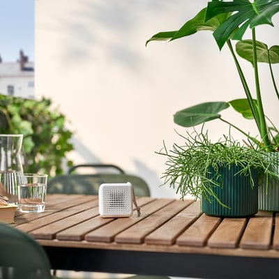 Small VAPPEBY white speaker on wooden table. Nearby, green potted plants and glasses. Perfect for outdoor use, compact and waterproof design.