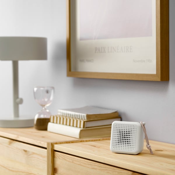 Small white portable speaker on a wooden surface near a lamp, books, and an hourglass.