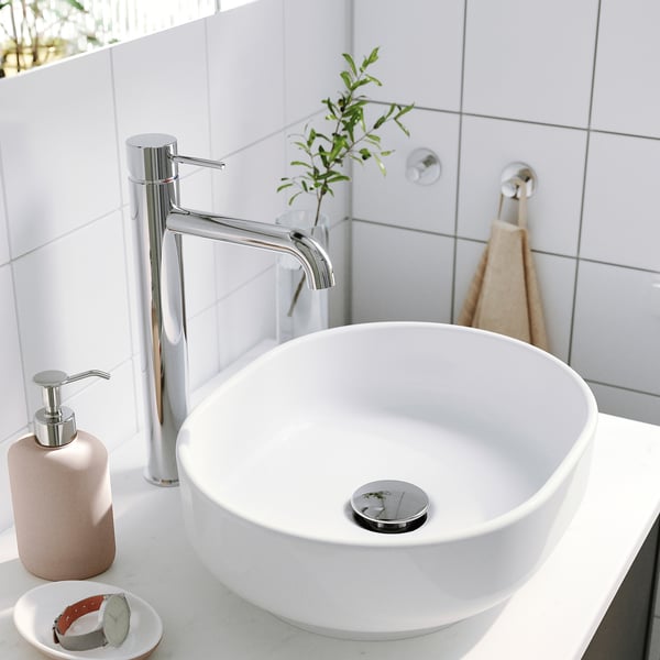 A modern bathroom sink with chrome tap, soap dispenser, plant & towel against white tiles.