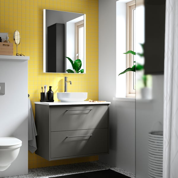 Bright bathroom with yellow tiles, white sink on grey cabinet, large mirror, small plant, and toilet. Grey rug on floor.