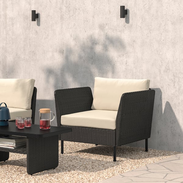 Modern black wicker outdoor lounge set with cushions, coffee table, wine glasses, and speaker, set against a grey concrete wall.