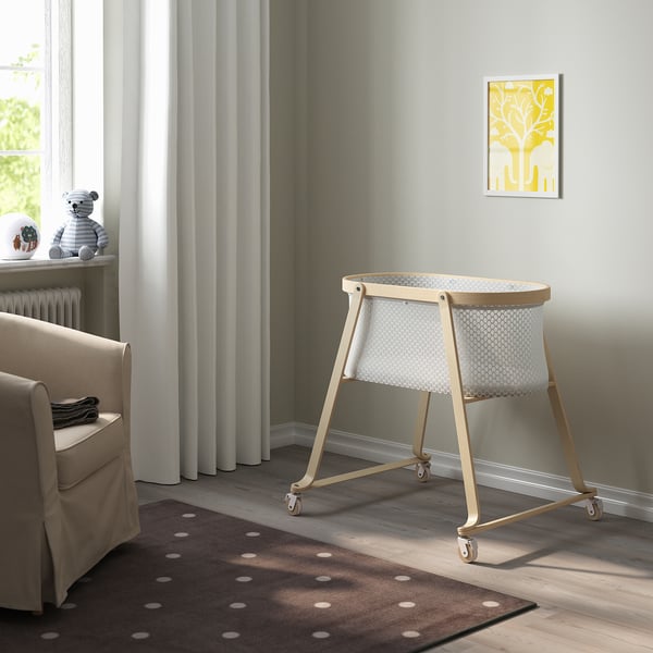 Modern nursery with ÄLSKVÄRD bassinet, polka-dot rug, beige chair, teddy bear, yellow frame.