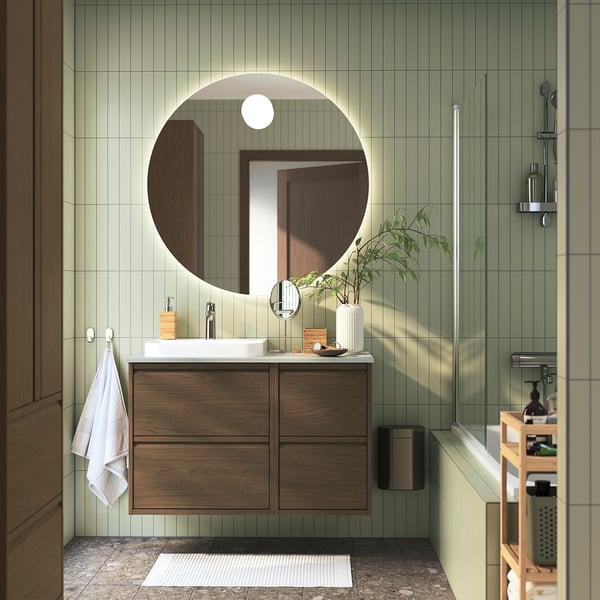 Modern bathroom with brown ÄNGSJÖN vanity, large mirror, green tiles, marble counter, towel, plant, partial tub.