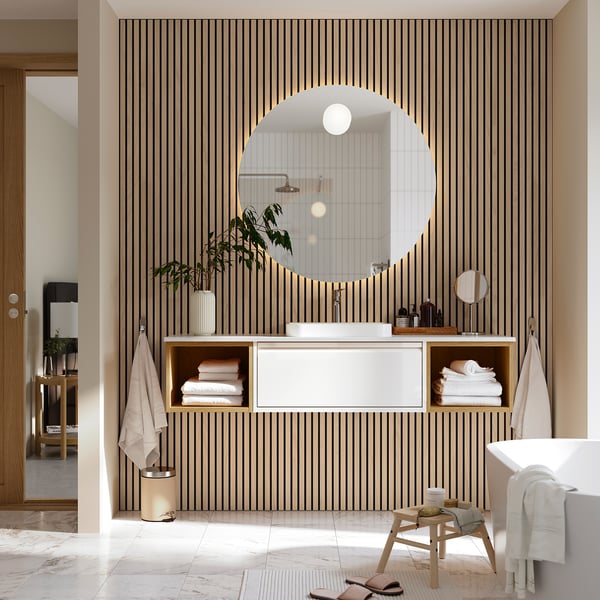 Modern bathroom with white floating cabinet, marble sink, and arched mirror against a striped wall. A wooden stool and towels are placed nearby.