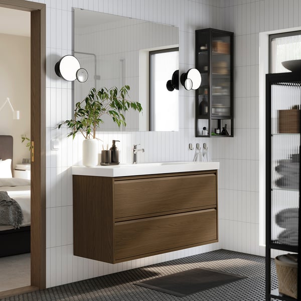 Modern bathroom with wall-mounted oak vanity, white counter, black accents, geometric mat, and plants.