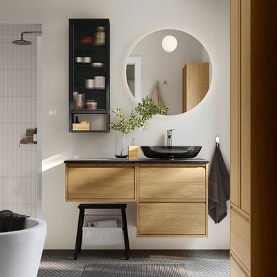 Modern bathroom with oak vanity, dark counter, glass basin, mirror, black towel, stool, magazine rack, rain shower, white tub.
