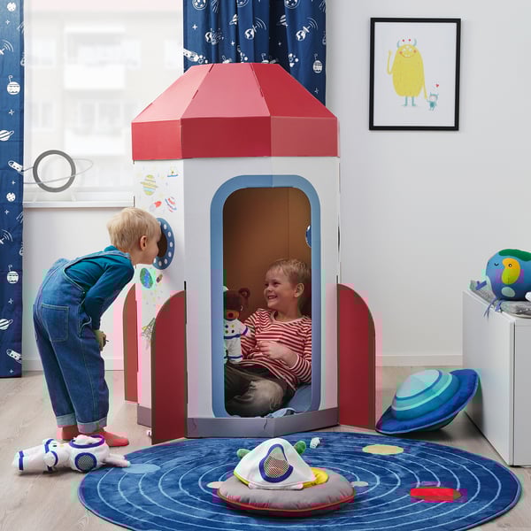 Two children play inside a colourful cardboard rocket ship, with a cuddly toy. A blue spaceship rug and plush alien are nearby.