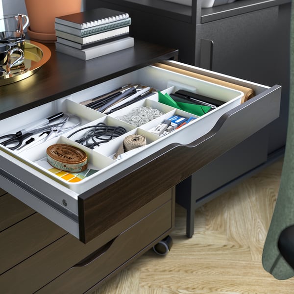 Organised desk drawer with compartments holding various small items like stationery, cables, and tape. Grey ALEX desk, wood floor, green chair.