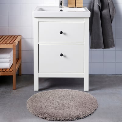 A white vanity with a sink and drawers, featuring a plush, beige ALMTJÄRN bath mat and a wooden bench with towels and a basket.