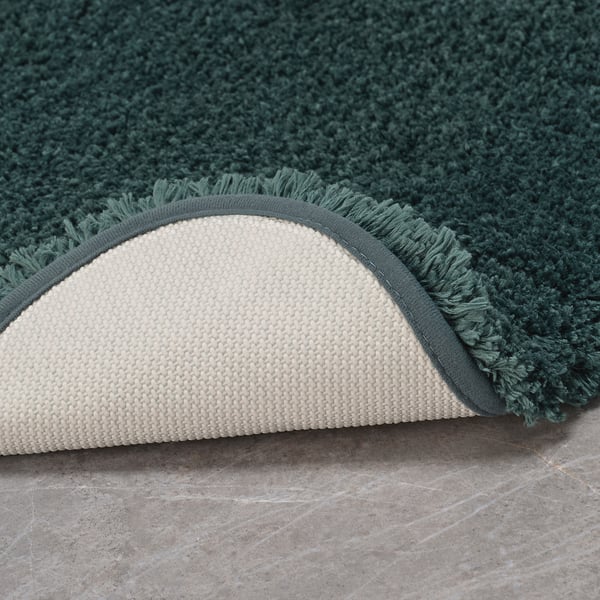 Green bath mat with fluffy texture and cream-colored non-slip backing, placed on a grey floor.