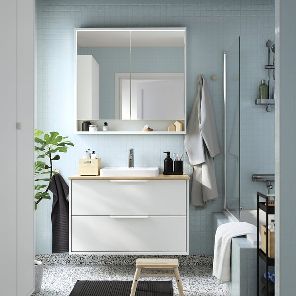 Modern bathroom with light blue tiles, sleek white cabinet, bamboo countertop, rectangular vanity mirror, towel rail, plant, and shower.