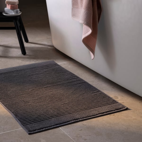 A dark grey bath mat named ALSTERN on a floor beside a tub. Its made of pure cotton, rectangular, and a ribbed texture.