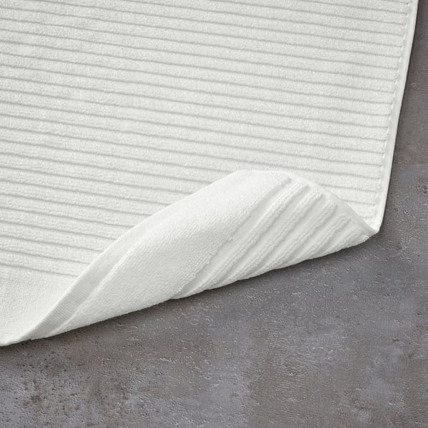 White bath mat with textured surface, laid out on dark grey floor. Designed for quick drying and high absorption.