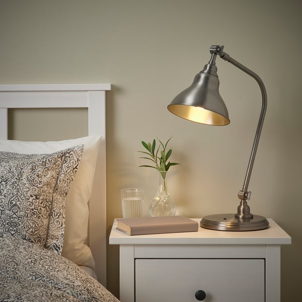 Pewter ANKARSPEL lamp on bedside table, illuminating book, adjustable arm against patterned pillows.