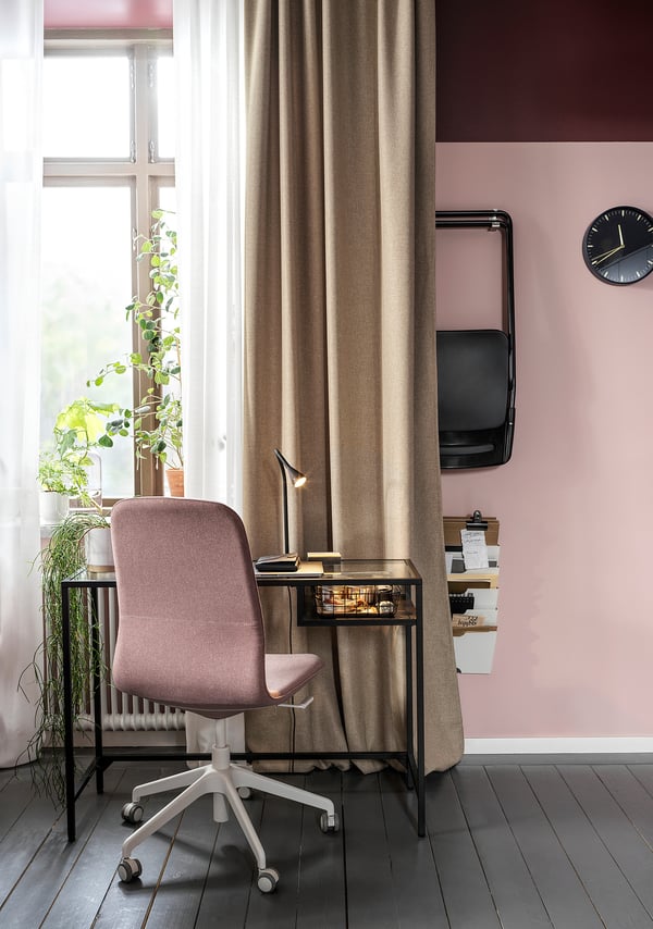 Cosy home office setup with pink ANNAKAJSA curtains, desk, chair, lamp, wall-mounted tv, clock, and plants. Dark floor, light walls.