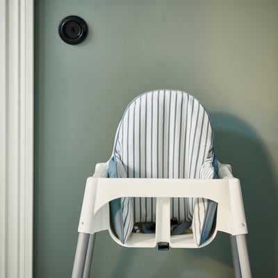 A white baby high chair with a striped cushion against a green wall, featuring a round power outlet above it.