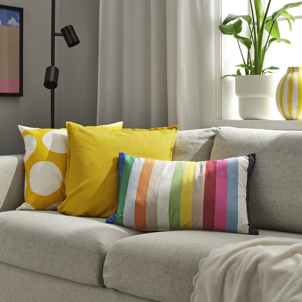 Bright yellow and multicoloured striped cushions on a grey couch, with a potted plant beside, near a window with curtains.
