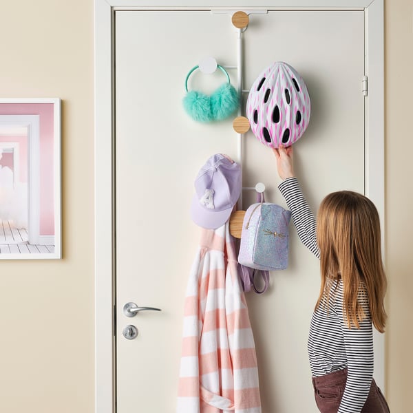 Image shows a child adjusting a pink helmet on a door-mounted coat hanger. The hanger holds a striped blanket, a hat, a backpack, and earmuffs.
