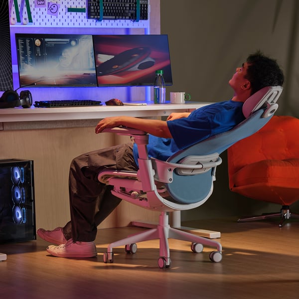 Gamer reclines in pink BÄSTBOLL chair, feet up, enjoying ergonomic comfort and mesh back with dual monitors and gear nearby.