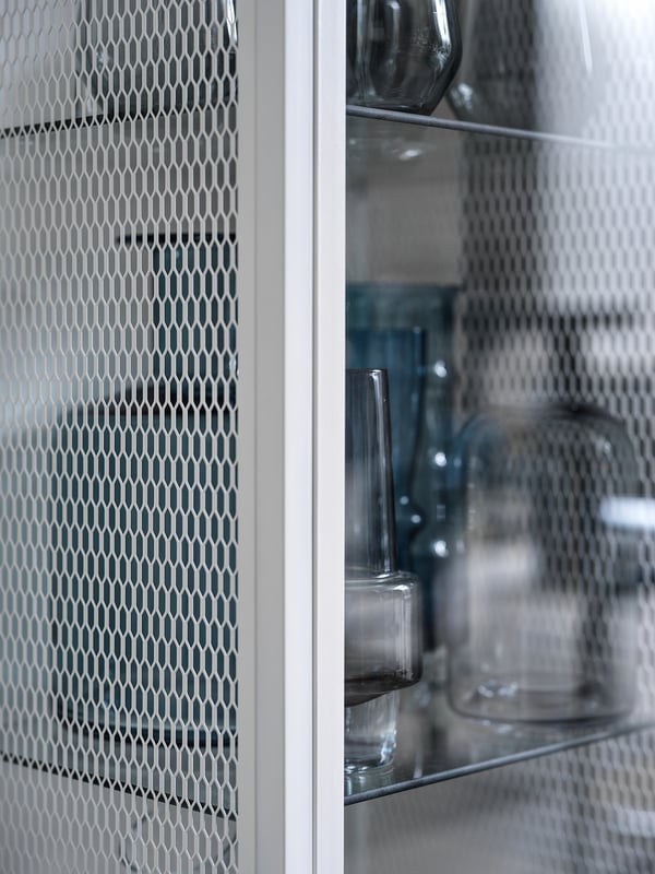 A stylish, white BAGGEBO display cabinet with a mesh and glass door. It holds clear glass vases on shelves.