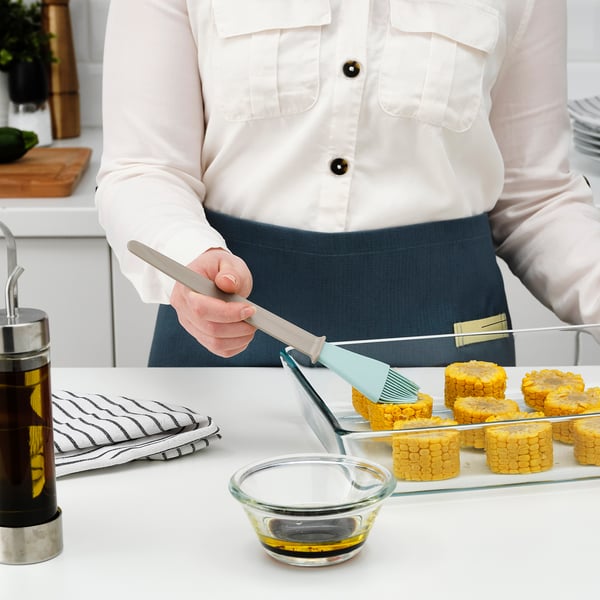Person in white shirt, holding a kitchen brush, preparing corn on the cob with oil in a bowl, and handling several pieces of corn.