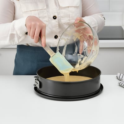 Person pouring batter from a spatula into a round cake pan. The spatula has a small support to avoid messes.