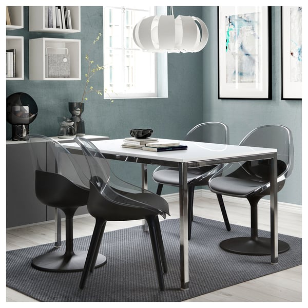 Modern dining set with clear chairs and white table, stylish light fixture, blue-grey wall, decorative art, and shelf setup.