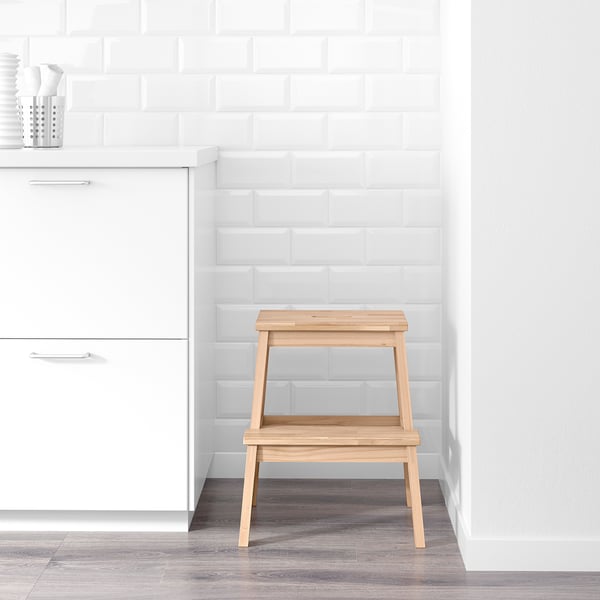 A wooden step stool with two steps in a white room, positioned against a white brick wall and next to white cabinetry.