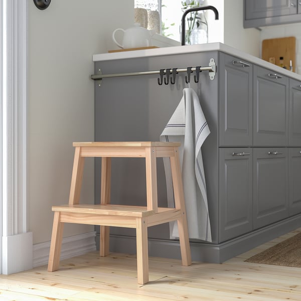 Wooden BEKVÄM step stool beside grey kitchen cabinets. Small, light-colored, stable.