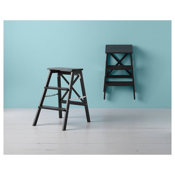 A black BEKVÄM foldable step ladder against a blue wall. It has three steps, is made of solid wood, and can be folded for space-saving.