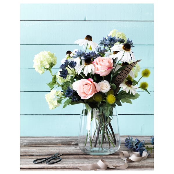 A clear glass vase with a bouquet of white, pink, and blue flowers sits on a wooden table against a turquoise wall.