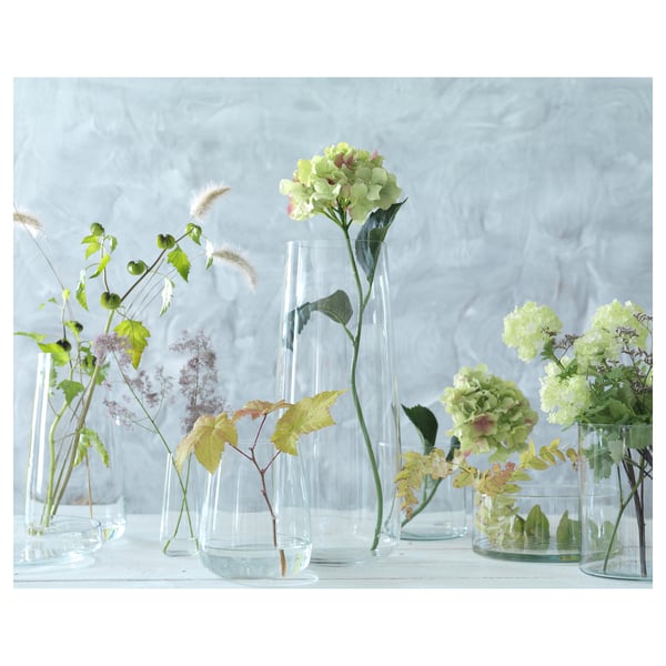 Clear glass vases in various shapes hold green flowers and foliage on a light surface.