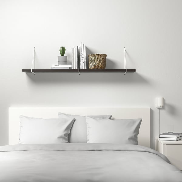 Modern white bedroom with BERGSHULT wall shelf. Shelf holds books, basket, and plant. Bed below with white bedding, bedside table with books.