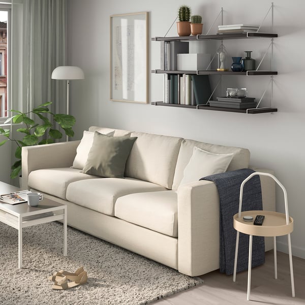 Modern living room with beige sofa, grey carpet, side table, stool, vases, art, shelves with books & plants.