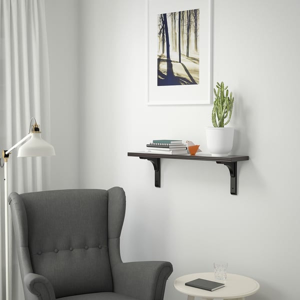 A cosy room with a grey chair and a BERGSHULT wall shelf. The shelf holds books, a plant, and a lamp. The shelf is smaller than the chair.