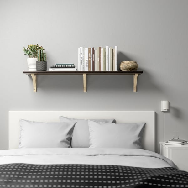 Modern bedroom with black shelf over bed displaying books, basket, cactus. Small bedside table with lamp and books.