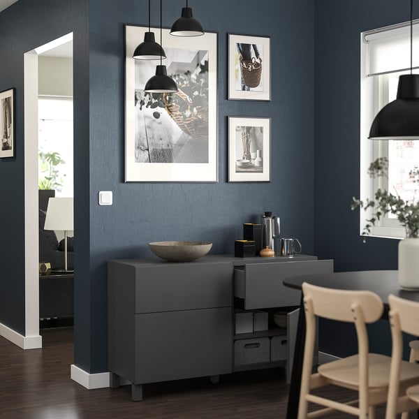 Modern living room with dark blue walls, grey BESTÅ storage unit, black pendant lights, framed photos, wooden chair, and plants.