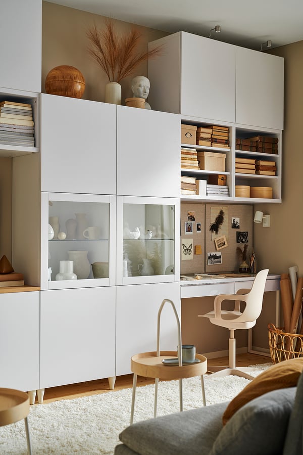Modern home office with white BESTÅ cabinets, glass doors, books, decorative items, chair, and small table with mug.