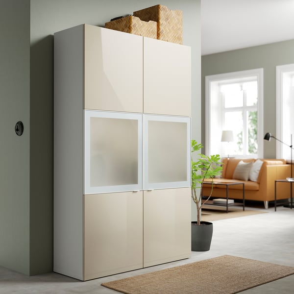 Corner storage unit, BESTÅ. Off-white, 4-door, glass panels. Tall, modern design. Plant & sofa beside.