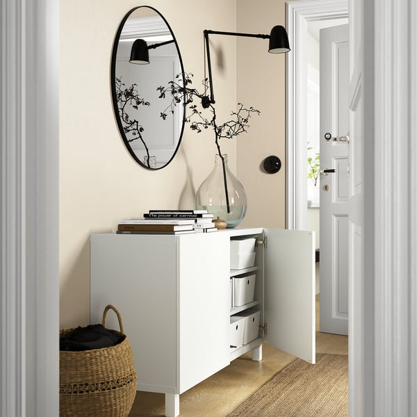 White BESTÅ sideboard with open door revealing storage boxes, beneath a round mirror and light fixture.