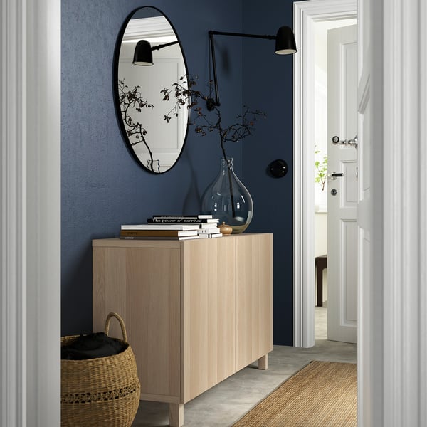 Light wood BESTÅ sideboard with two doors, dark blue wall, black oval mirror, stacked books, vase, basket, bright room peek, black wall lamps.