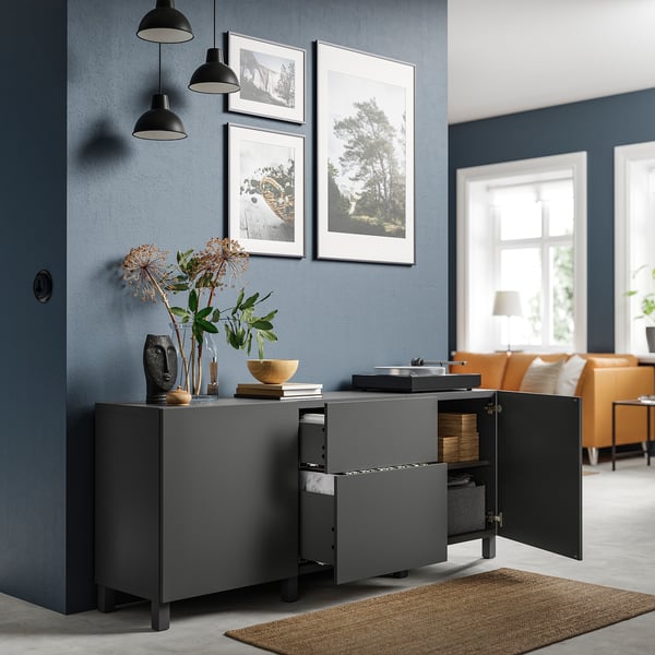 Modern grey BESTÅ storage unit with open drawers and cabinet, black handles, steel hinges, and decorative items on top, against blue wall with photos.