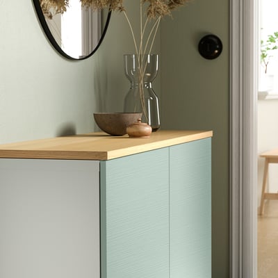 Modern BESTÅ sideboard in light blue with oak top, decorated with glass vase, bowl, and jar, with mirror, door, and curtain.