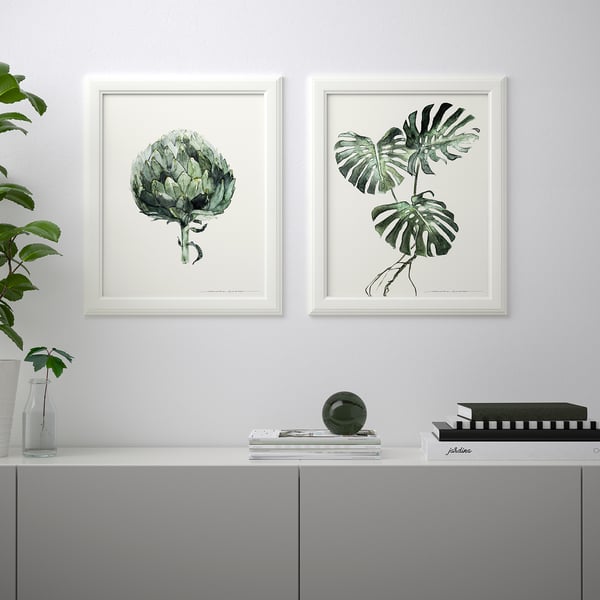 Two framed botanical prints hang above a grey sideboard, which holds a plant, vase, book, magazine, and decorative sphere.