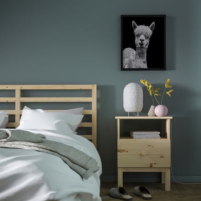 Cosy bedroom with light wood bed, white bedding, blue wall, lamp, flowers, books, alpaca poster, grey slippers.