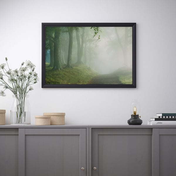 A framed nature scene above a grey cabinet, with flowers, boxes, and a lamp.