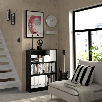 Cozy room with black BILLY bookcase, lamp, headphones, guitar, and clock. Large window, striped cushion, and bright natural light.