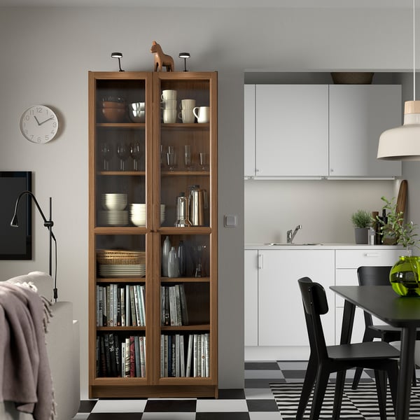 BILLY bookcase with glass doors, filled with items, between a dining area and kitchen in a modern home.